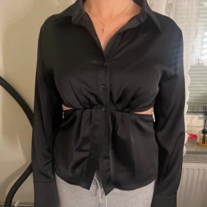 Svart blus från Gina Tricot - Snygg svart blus från Gina Tricot med knappar framtill och en unik design med öppna sidor. Blusen har långa ärmar och en klassisk krage, perfekt för en stilren look.