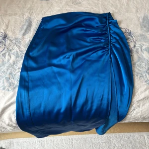 Blå satin kjol från Zara - Snygg blå satin kjol från Zara med en elegant omlott-design. Kjolen har en smickrande passform och en subtil drapering på sidan som ger en fin detalj. Perfekt för att ge en touch av lyx till din outfit.