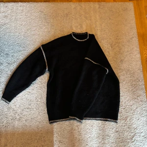 Svart stickad tröja med mönstrade kanter - Säljer en svart stickad tröja med vita mönstrade kanter vid hals, ärmslut och nederkant. Tröjan har långa ärmar och en klassisk passform. Perfekt för kyligare dagar!