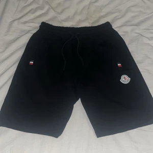 Svarta shorts från Moncler - Snygga svarta shorts från Moncler med elastisk midja och snörning. Shortsen har Moncler-logga och små detaljer i rött, vitt och blått. Perfekta för en stilren look.