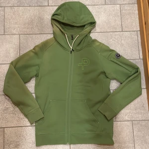Grön huvtröja från Pelle P - Säljer en snygg grön huvtröja från Pelle P med dragkedja och en stilren logga på ärmen. Tröjan har långa ärmar och en praktisk huva. Perfekt för en avslappnad stil. Knappt använd, men har blivit solblekt på vissa ställen.