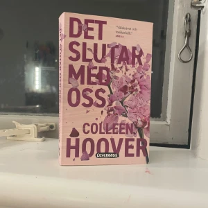 Det slutar med oss - Boken 'Det slutar med oss' av Colleen Hoover, utgiven av Lovereads. Omslaget är rosa med blommotiv och stora lila bokstäver. En populär roman som berör djupa känslor och relationer.