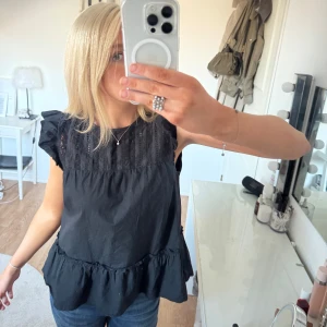 Svart  blus med volang - Säljer en stilren svart blus med volangdetaljer vid ärmarna och nederkanten. Blusen är ärmlös och har en luftig passform, perfekt för både vardag och festliga tillfällen.