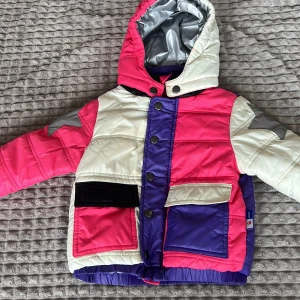 MOLO jacka - Säljer en cool och färgglad quiltad jacka med huva. Jackan har en mix av rosa, lila och vit med en grå stjärna på ärmen. Den har knappar och dragkedja framtill samt praktiska fickor.