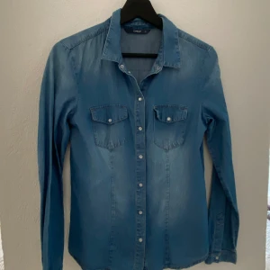 Blå jeansskjorta från Only i stretch material - Snygg blå jeansskjorta från Only med långa ärmar och knappar framtill. Skjortan har två bröstfickor med knappdetaljer. Perfekt för en avslappnad stil.