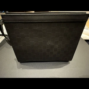 Svart axelväska med rutigt mönster - Snygg svart axelväska med ett diskret rutigt mönster. Väskan har en stilren design och är perfekt för att bära dina dagliga nödvändigheter. Den har en axelrem för enkel bärning.