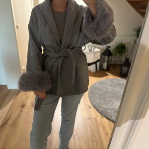Grå kappa med fuskpälsdetaljer - Säljer en elegant och helt ny grå kappa i storlek S. Pälsdetaljer finns vid ärmsluten och är avtagbara, så du kan välja att bära kappan med eller utan dem (se bilder).  Pris kan diskuteras. Skicka gärna ett meddelande vid intresse! ❤️