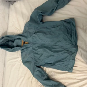 Blå vindjacka från Tiger of Sweden - Snygg blå vindjacka från Tiger of Sweden i storlek S. Jackan har en dragkedja framtill och en huva. Yttermaterialet är gjort av nylon och fodret är av bomull, vilket gör den både lätt och bekväm. Perfekt för blåsiga dagar!