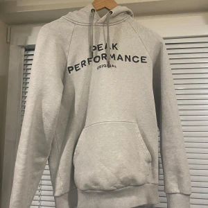 Grå hoodie från Peak Performance - Säljer en stilren grå hoodie från Peak Performance med svart text på bröstet. Tröjan har en klassisk känguruficka och justerbar huva med snören. Perfekt för en avslappnad look.