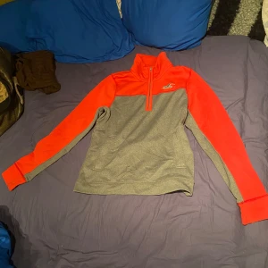 Grå och röd tröja från Hollister - Säljer en grå och röd tröja från Hollister med dragkedja vid halsen. Tröjan har långa ärmar och en sportig look, perfekt för kyligare dagar. Hollister-loggan syns på bröstet och ryggen.