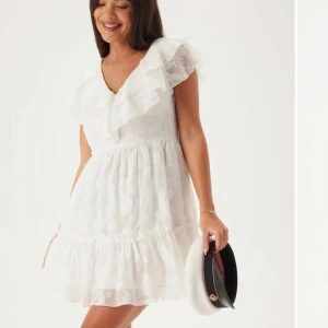 Vit klänning med volangdetaljer - En söt, vit klänning med volangdetaljer och v-ringning. Klänningen har en kort längd och är perfekt för somriga dagar och studenten. Den är ärmlös med en luftig design. Ny och oanvänd.