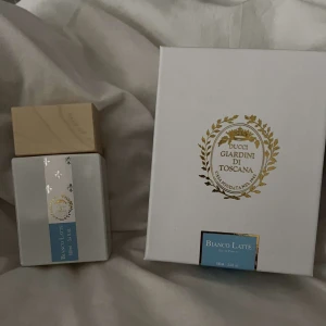 Bianco Latte parfym från Giardini di Toscana - Elegant parfymflaska med en stilren design från Giardini di Toscana. Flaskan är vit med en beige kork och har en dekorativ etikett i blått och guld. Förpackningen är vit med gyllene detaljer och logotyp. Perfekt för den som söker en sofistikerad doftupplevelse.