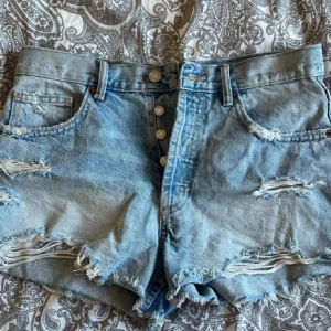 Blå jeansshorts från Zara - Säljer ett par coola blå jeansshorts från Zara med slitna detaljer och fransar. Shortsen har knappar framtill. Perfekta för en avslappnad stil!