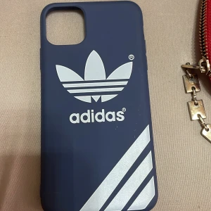 Blå mobilskal från Adidas - Snyggt blått mobilskal från Adidas med det ikoniska trefoil-loggan i vitt. Skalet har även de klassiska tre ränderna i vitt på framsidan. Perfekt för att skydda din telefon med stil.