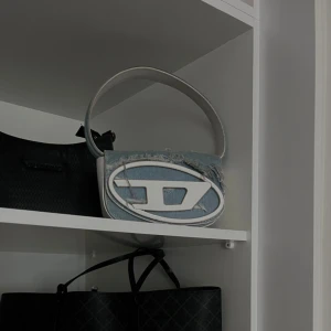 disel väska  - Säljer en unik jeansväska med en stor logotyp på framsidan. Väskan har en rund form och ett bekvämt handtag. Perfekt för att ge en cool touch till din outfit. 
