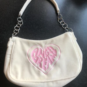 Vit axelväska med rosa text - Säljer en söt vit axelväska med texten 'Angel Dream' i rosa på framsidan. Väskan har en kedjedetalj på axelremmen och är perfekt för att ge en touch av stil till din outfit. Den är tillverkad i ett mjukt material och har en dragkedja för säker förvaring.