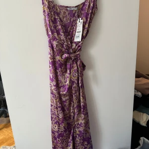 Lila mönstrad klänning från Zara - Säljer en lila klänning från Zara med ett vackert mönster i guld och beige. Klänningen har en omlott-design med knytning i midjan och är ärmlös. Perfekt för sommarens festligheter!