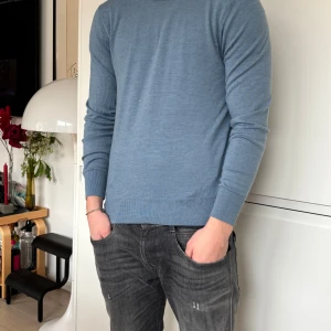 Blå stickad tröja - Säljer en stilren blå stickad tröja med rund halsringning. Tröjan har långa ärmar och ribbade muddar vid ärmslut och nederkant. Perfekt för en avslappnad look.