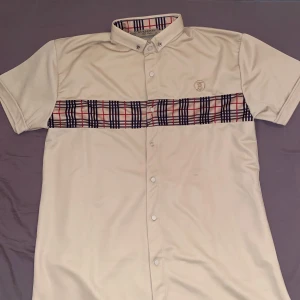 Beige kortärmad skjorta från Burberry - Säljer en stilren beige kortärmad skjorta från Burberry med klassiskt rutmönster över bröstet och insidan av kragen. Skjortan har knappar framtill och en broderad logotyp på bröstet. Perfekt för en elegant look.