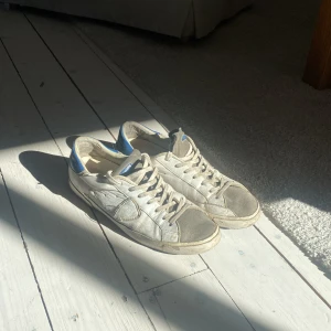 Vita sneakers från Philippe Model - Säljer ett par vita sneakers från Philippe Model med blå detaljer. Skorna har snörning och en klassisk design med en bekväm passform. Perfekta för en avslappnad stil. De är ganska slitna och lite smutsiga. Men inget man inte kan tvätta bort. Allt OG följer med: dustbag, boxen och kvitto!