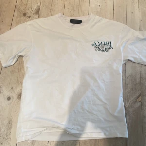 Vit t-shirt från Amiri  - Säljer en vit t-shirt från Amiri med ett snyggt blommigt tryck i blått på både framsidan och baksidan. Perfekt för en stilren och avslappnad look. T-shirten har korta ärmar och en normal passform.