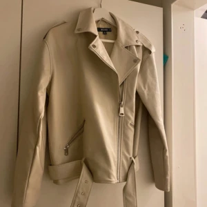 Beige skinnjacka från Missguided - Snygg beige skinnjacka från Missguided med dragkedja och bälte i midjan. Jackan har långa ärmar och en klassisk krage med knappar. Perfekt för en stilren look.