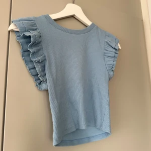 Ljusblå ribbad topp med volangärmar - Snygg ljusblå ribbad topp med volangdetaljer på ärmarna. Toppen har en rund halsringning och är tillverkad i ett mjukt material. Perfekt får vår och sommar💙🦋, liten i storlek!