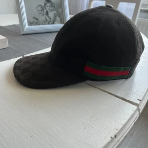 Svart keps från Gucci - Snygg svart keps från Gucci med ett diskret mönster och klassiska röd-gröna ränder på sidan. Kepsen har en justerbar rem baktill för perfekt passform. Perfekt för att ge din outfit en lyxig touch.