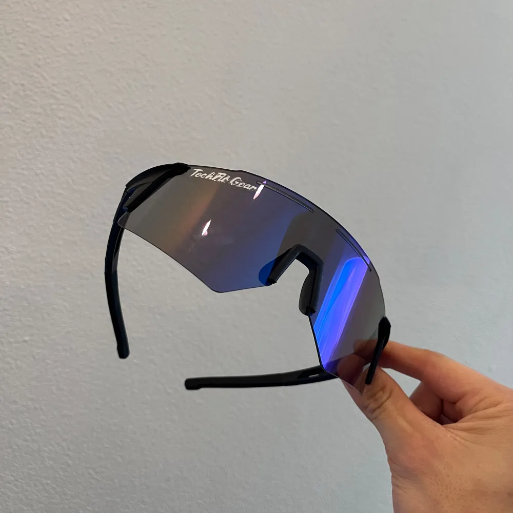 Snygga sportglasögon från TechFit Gear med en futuristisk design. De har en spegelblå lins och svarta bågar, perfekt för utomhusaktiviteter. Glasögonen har en omslutande passform som ger bra skydd mot solen.. Asusteet.