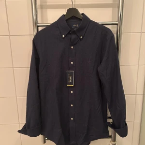 Mörkblå skjorta från Polo Ralph Lauren - Snygg mörkblå skjorta från Polo Ralph Lauren med klassisk button-down krage och vita knappar. Skjortan är i custom fit och tillverkad i bomull, perfekt för en stilren look. Diskret logga på bröstet och långärmad modell.