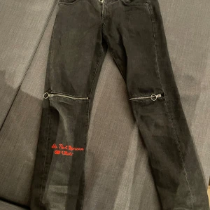 Svarta jeansbyxor från Off-White - Snygga svarta jeansbyxor från Off-White med unika dragkedjedetaljer vid knäna och röd broderad text på benet. Perfekta för dig som vill ha en edgy stil. Byxorna har en klassisk femficksdesign och är tillverkade i slitstarkt denim. Sitter större än S