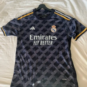 Real madrid tröja - Säljer en snygg svart Real Madrid fotbollströja från Adidas med ett unikt mönster och klubbens emblem på bröstet. Tröjan har korta ärmar och gula detaljer på axlarna. Perfekt för alla fotbollsfans! ⚽️lite stor på mig som är 183