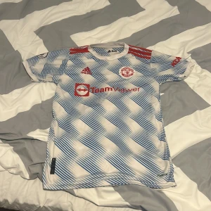 Man United tröja, Vit och blå - Säljer en vit och blå Manchester United med röda detaljer. Tröjan har korta ärmar och ett randigt mönster. Perfekt för fotbollsfans!