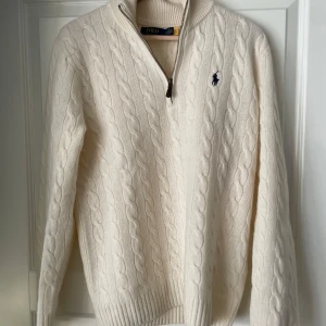 Vit stickad tröja från Ralph Lauren - Säljer en stilren vit stickad tröja från Ralph Lauren med kabelstickat mönster och en liten broderad logga på bröstet. Tröjan har en dragkedja vid halsen och långa ärmar. Perfekt för kyligare dagar! Nypris - 2 795