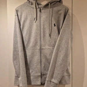 Grå hoodie från Ralph Lauren - Säljer en stilren grå hoodie från Ralph Lauren med dragkedja och klassisk logga på bröstet. Tröjan har långa ärmar och en bekväm huva med snören. Perfekt för en avslappnad look.