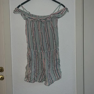 Randig jumpsuit från H&M - Säljer en söt randig jumpsuit från H&M i ljusa färger med offshoulder-design och volangdetaljer. Det är även shorts i denna fina jumpsuiten Perfekt för varma sommardagar. Den har smala axelband och en bekväm passform.