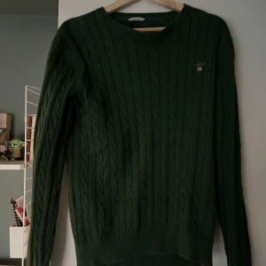 Grön kabelstickad tröja från GANT - Säljer en snygg grön kabelstickad tröja från GANT. Tröjan har en klassisk design med rund hals och långa ärmar. Perfekt för kyligare dagar och ger en stilren look. Passar bra till både jeans och chinos.