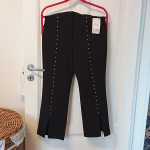 Svarta byxor med nitar - Snygga svarta byxor med nitar längs benen och slitsar vid anklarna. Perfekta för en trendig look. Byxorna har en elegant design och passar bra till både vardag och festligare tillfällen. Storlek M