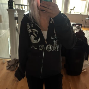 Svart hoodie med dragkedja från Aim for the Moon - Säljer en svart hoodie med dragkedja från Aim for the Moon. Den har ett coolt tryck med glittriga detaljer på framsidan. Perfekt för en avslappnad stil. Hör av dig vid intresse!