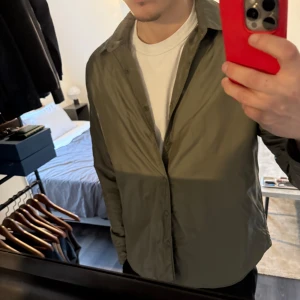 Jacka/overshirt från  Aspesi - Säljer en stilren oliv grön overshirt från italienska Aspesi i nylon taft, som är både mjuk och vattenavvisande. Overshirten har tryckknappar fram och vid ärmslut samt en rundad fåll. Perfekt för att hålla värmen med sin Thermore polyesterstoppning. Passform M/L