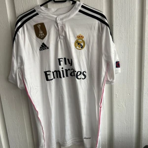 Vit Real Madrid fotbollströja - Säljer en vit Real Madrid fotbollströja från Adidas med korta ärmar. Tröjan har svarta och rosa detaljer samt klubbens emblem och sponsorlogga på framsidan. Baksidan pryds av namnet 'Ronaldo' och nummer 7. Perfekt för fans av laget!