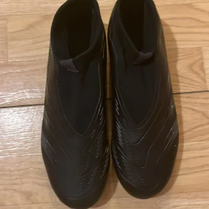 Svarta fotbollsskor från Adidas - Säljer ett par svarta Adidas Predator fotbollsskor. De har en strömlinjeformad design med diskreta mönster och är tillverkade för att ge bra grepp och kontroll på planen. Perfekta för fotbollsspelare som vill ha stil och funktionalitet. Och de e oanvända o helt nya.