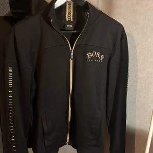 Svart tröja från Hugo Boss - Snygg svart tracksuit tröja från Hugo Boss med dragkedja och ribbad krage. Tröjan har långa ärmar med stilrena detaljer längs sidorna. Perfekt för en stilren eller sportig look. Strlk L passar m och s (Nyskick)