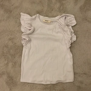 Vit ärmlös topp från Uniq Studio - Säljer en söt vit ärmlös topp från Uniq Studio med volangdetaljer längs axlarna. Perfekt för en stilren och chic look. Toppen är ribbad och har en mjuk och bekväm passform.