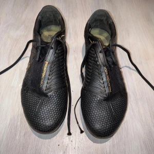 Svarta fotbollsskor från Nike - Snygga svarta fotbollsskor från Nike med gulddetaljer och snörning. Skorna har en texturerad yta för bättre grepp och kontroll på planen. Perfekta för fotbollsträning och matcher.