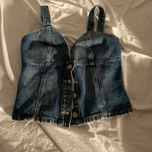 Jeanskorsett med knappar - Säljer en snygg jeanskorsett med knappar framtill och  axelbanden är ihopsydda så den inte skulle vara så hänging vilket man kan se på ryggen men det är inte synligt. Toppen har en smickrande passform och elastisk rygg för extra komfort. Perfekt för att ge en cool touch till din outfit på fest❤️❤️