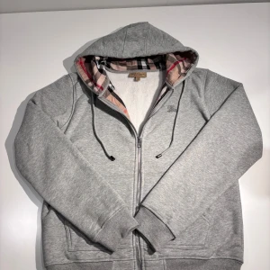 Grå hoodie från Burberry - Säljer en stilren grå hoodie från Burberry med dragkedja och klassiskt rutigt mönster i huvan. Tröjan har en broderad logga på bröstet och är tillverkad i mjuk bomull. Perfekt för en avslappnad look.