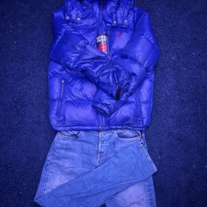 Blå dunjacka från Polo Ralph Lauren - Säljer en snygg blå dunjacka från Polo Ralph Lauren. Jackan har en dragkedja framtill och en praktisk huva. Perfekt för kyliga dagar och håller dig varm och stilren. Den har långa ärmar och en klassisk design med diskret logotyp på bröstet.