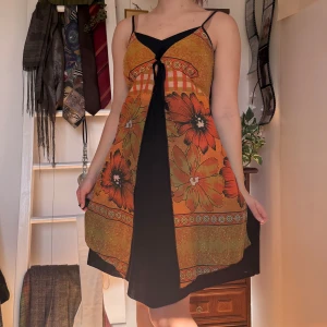 Vintage Orange blommig klänning    - Säljer en färgglad klänning med blommönster i orange och svart. Klänningen har smala axelband och en v-ringad design med knytning framtill. Perfekt för sommarens varma dagar! Köpt secondhand men har inte använt särskilt mycket. 