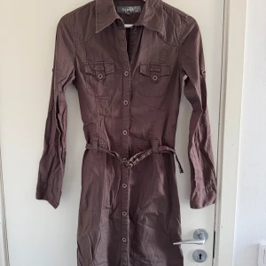 Brun klänning från Clara - Säljer en brun klänning från Clara med långa ärmar och knappar framtill. Klänningen har en bälte i midjan för en snygg passform och två bröstfickor med knappar. (Katt och kanin finns i hemmet) 